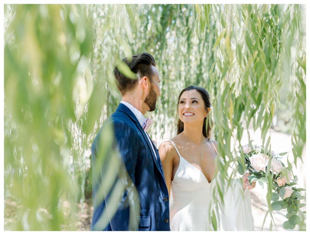 A bride and groom smile at each other under the willow trees at TERRA MIA Vineyards a wine country wedding venue in Paso Robles, California