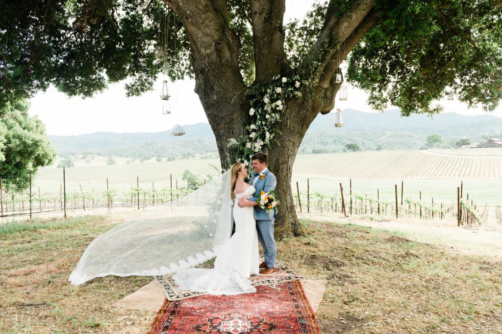 Bride and groom pose under giant oak tree at Oyster Ridge Vineyards for whimsical boho wedding photo in Paso Robles.