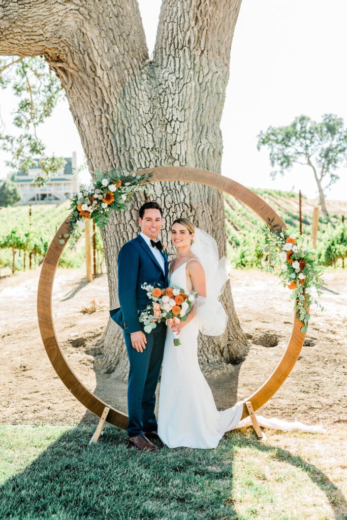 A bride and groom in wedding day attire stand before their circular arch underneath the giant oak tree at Ellas Vineyard.