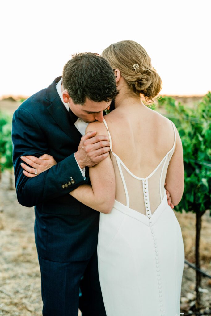 A groom gives the bride, a romantic kiss on her shoulder while they stand in the sunset in the vineyards at Ella's Vineyard on their wedding day. 