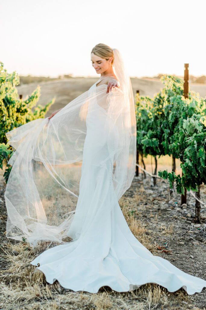 A bride plays whimsically with her wedding veil in the vineyards at Ellas Vineyard on her wedding day during sunset. 