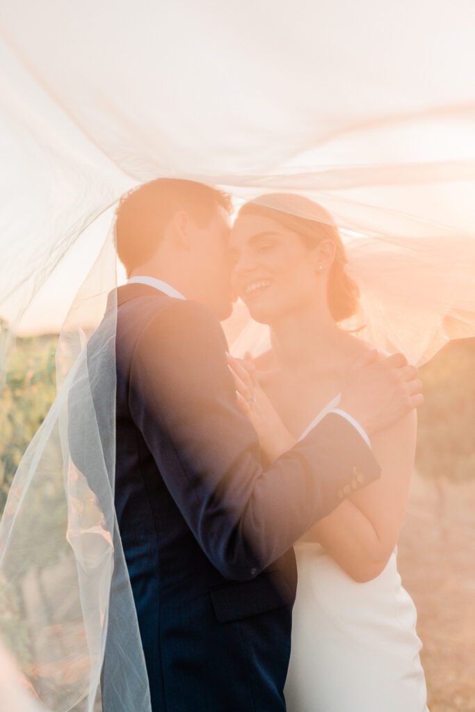 A bride and groom, laughing underneath the wedding veil during golden hour photos at Ellas Vineyard in Paso Robles.