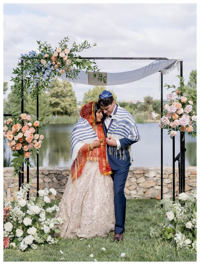 Bride and groom sharing first kiss under floral chuppah at Jewish-Hindu fusion wedding ceremony in Paso Robles, photographed by Leana Myra Photography