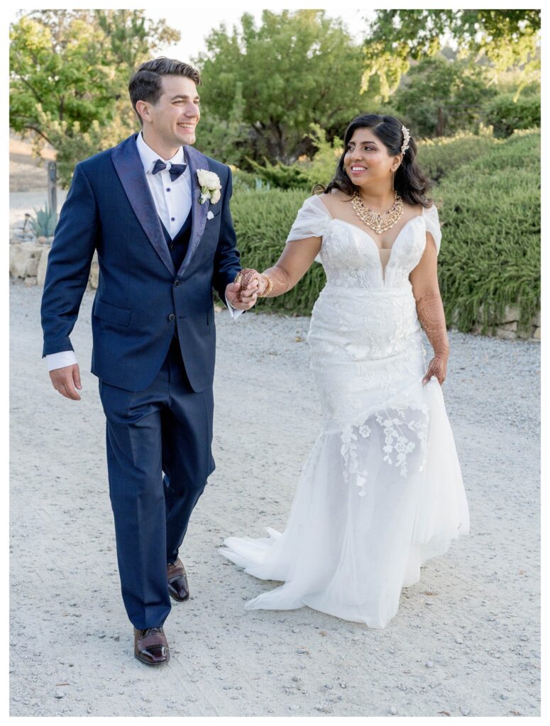 Joyful bride and groom walking hand-in-hand in vineyard setting at Paso Robles wedding captured by Leana Myra Photography.