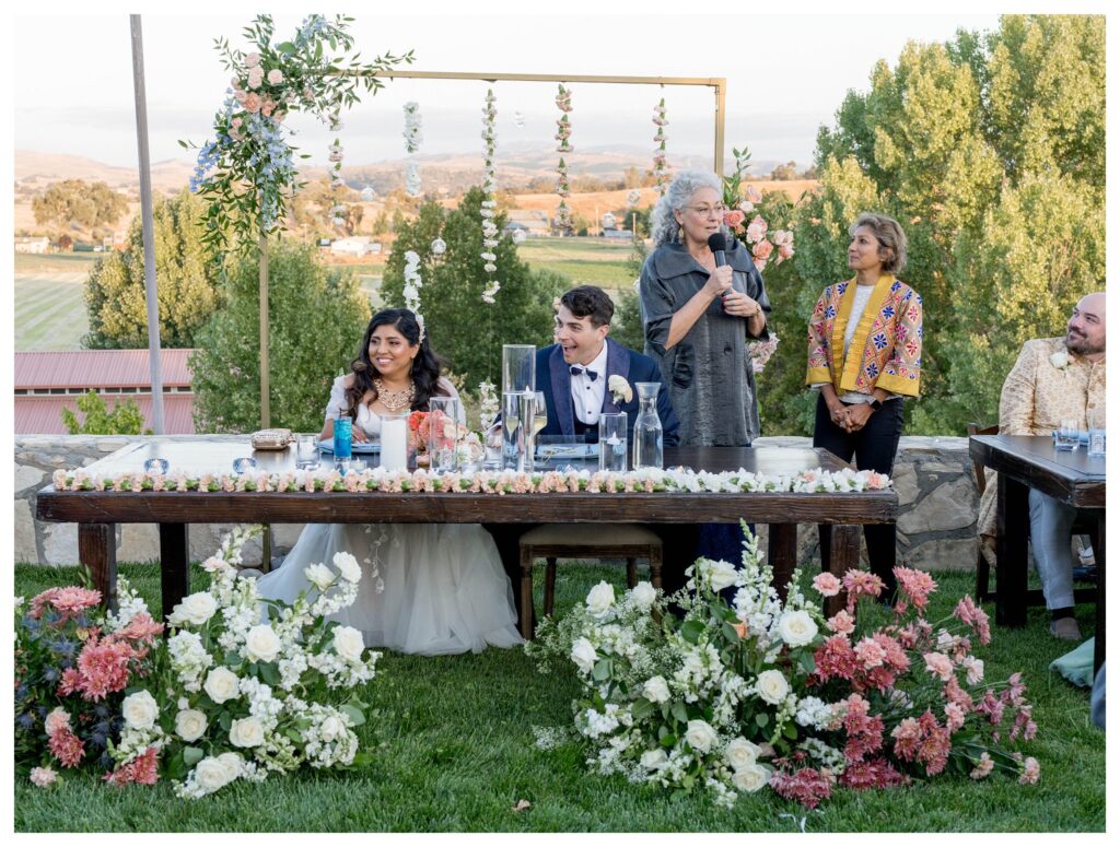 Bride and groom smiling at sweetheart table during heartfelt speeches, surrounded by lush florals at Terra Mia Vineyards in Paso Robles, captured by Leana Myra Photography.