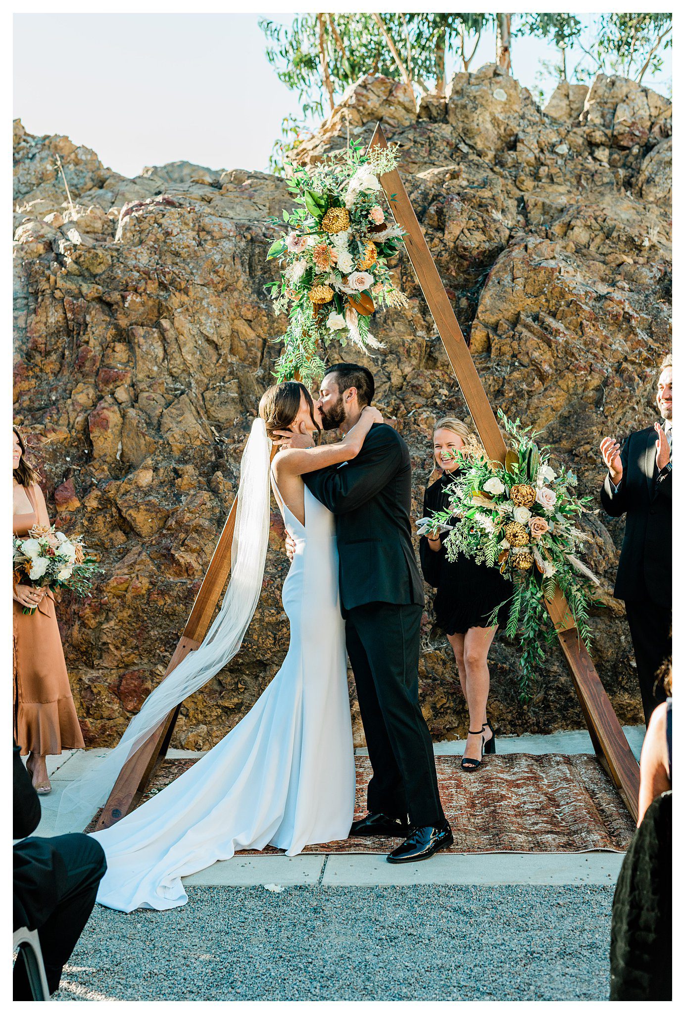 Slo brew Rock wedding ceremony, bride and groom kissing in front of a boho style triangle ceremony arch and warm fall colors in san luis obispo.