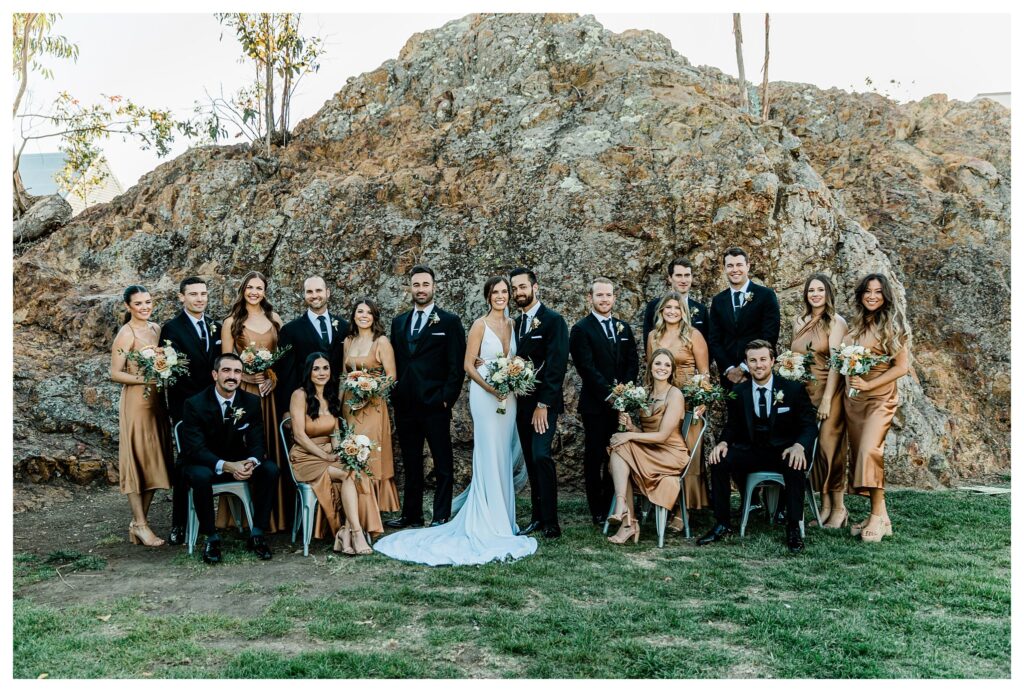 Bridesmaids and groomsmen in warm fall colors pose together in front of the rock at Slo Brew Rock, a san luis Obispo vineyard wedding venue.