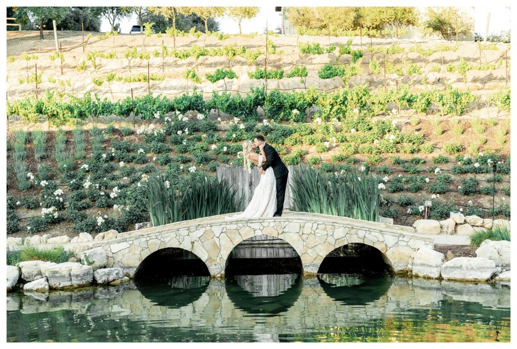 A bride and groom kiss romantically on the bridge at Terra mIa Vineyards next to the sparkling pond in the garden filled with white roses.