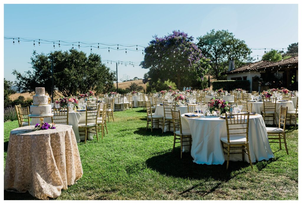A bright and colorful wedding reception at the Casitas Estate a wedding venue in San Luis Obispo with a lush garden overflowing with flowers. Tables with pink floral arrangements are surrounded by gold chairs and a luxury wedding cake. 