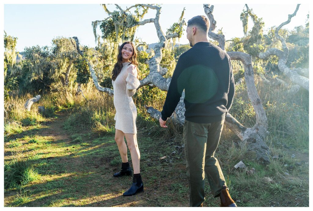 Woman beckons to man playfully during their candid engagement photos in a magical and dreamy forest near San Luis Obispo.