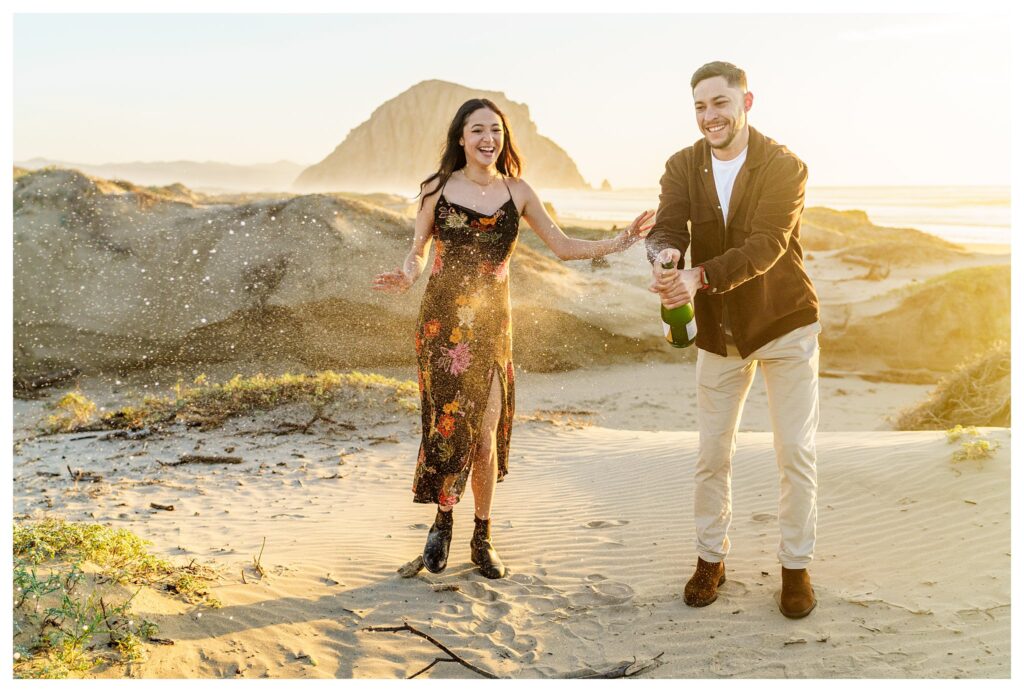 An engaged couple laughs playfully as they pop a bottle of champagne at their engagement session on the sand dunes in San Luis Obispo.