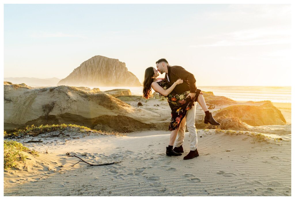 A woman and a man stand on a Sandune kissing passionately with a man dipping the woman back as you can see the Morro Bay rock in the distance, a beautiful and romantic scene of an engaged couple at Sunset in Morro Bay.
