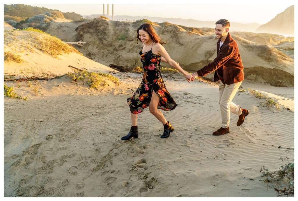 A woman and a man run over Sandunes during their Morro Bay engagement session, laughing, whimsically, and playfully.