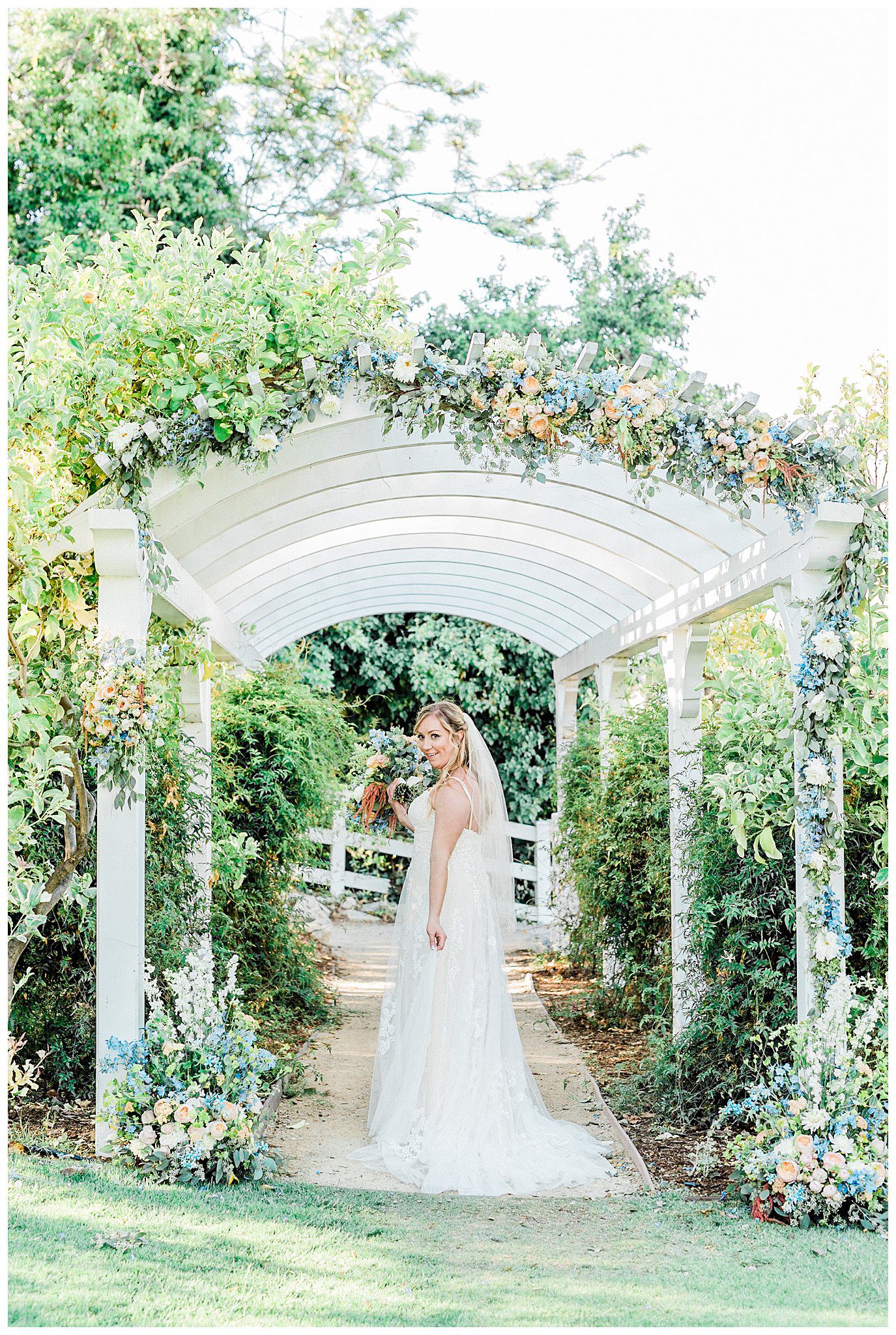 A bride under the arbor in the secret garden at the Madonna Inn, wearing her wedding dress and holding a bouquet of spring flowers.