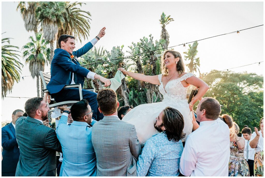 A bride and groom laugh as they are lifted up on chairs during a traditional Jewish dance at their wedding at the Santa Barbara zoo.