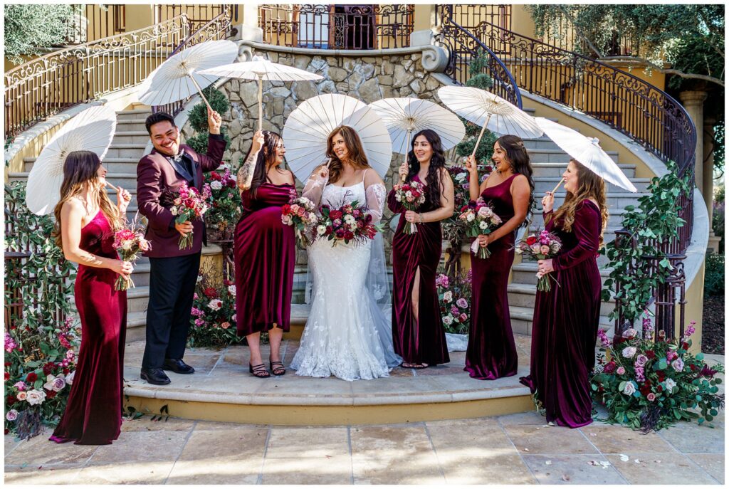 Bridesmaids during a luxury wedding, wearing velvet red dresses and holding umbrellas in Paso Robles, California.