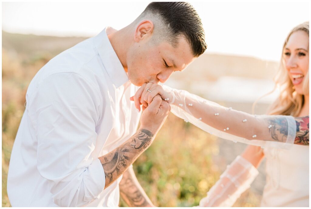 A groom kisses a brides hand on the beach for a picture.
