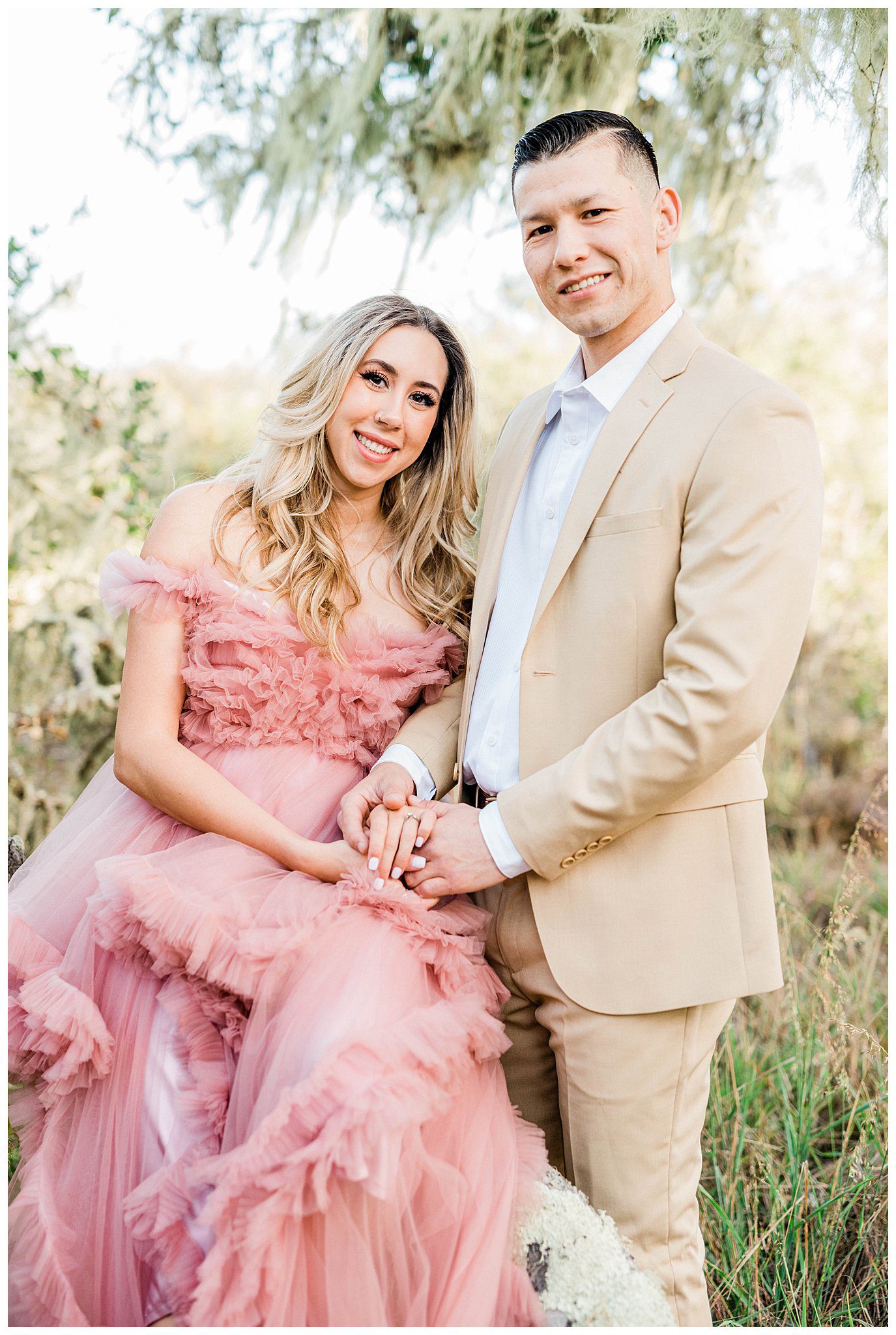 An engaged couple wearing a tan suite and a fluffy pink whimsical tulle dress smile at the camera during their central coast engagement session in the Los osos Oaks reserve. 