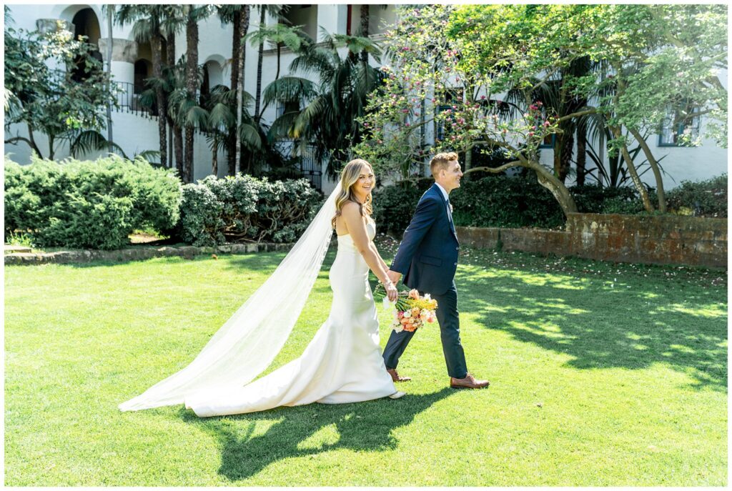 A bride and groom stroll across the lawn and wedding attire at the Santa Barbara courthouse during their elopement.