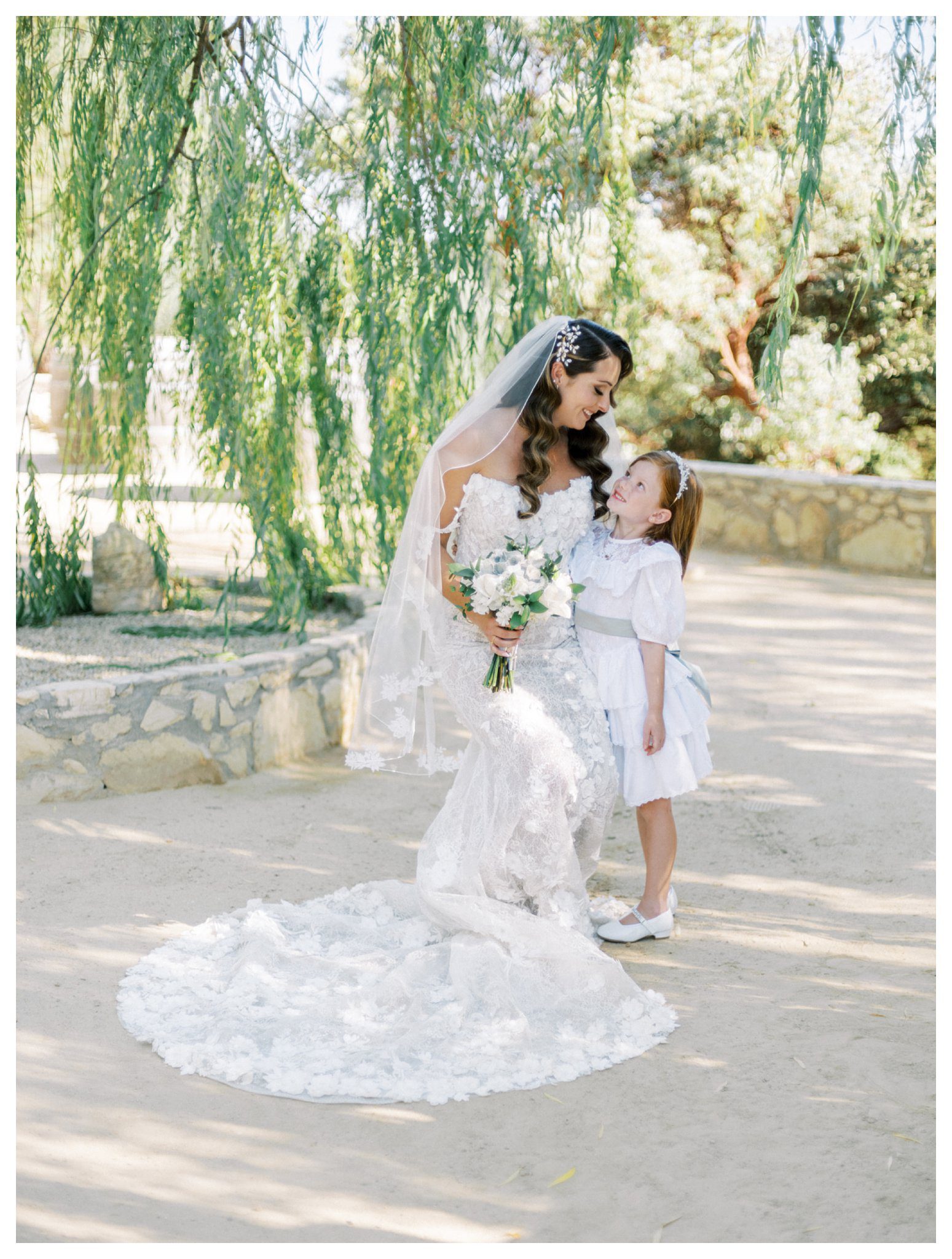 Bride kneeling with flower girl under willow tree